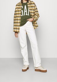 Camisa de cuadros amarilla y gris, camisa verde oliva con texto blanco, pantalones blancos, zapatillas con plataforma color beige. Tela suave, ajuste casual, look en capas.