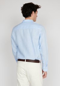Chemise à manches longues bleu clair en tissu doux, avec un empiècement au dos, des poignets à bouton et une texture lisse. Portée avec un pantalon de couleur claire.