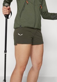 Pantalones cortos de senderismo verdes con un material ligero y elástico; cuenta con un logo blanco en el lateral y una abertura lateral para mayor movilidad.