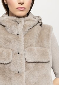 Frau trägt eine beige Kunstpelzweste mit zwei Fronttaschen über einem hellbeigen Pullover, der auf Mund- und Taillenhöhe zugeschnitten ist.