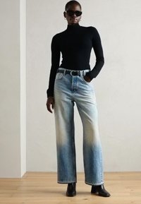Pull à col roulé noir à manches longues, jeans large bleu clair avec un effet dégradé et une ceinture noire. Porté avec des bottines noires à talons et des lunettes de soleil.