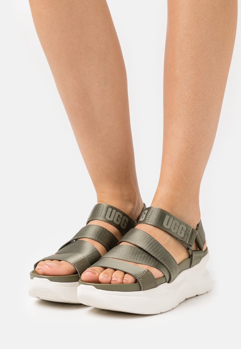 Olivově zelené sandály se širokými texturovanými pásky a bílými klínovými podrážkami. Obsahují branding UGG na pásech a mají otevřenou špičku.