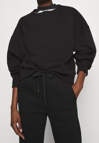 Sudadera negra de gran tamaño con hombros caídos, cuello redondo y puños acanalados; combinada con pantalones de chándal negros a juego que cuentan con un cordón.