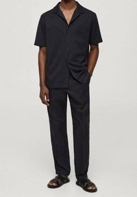 Uomo che indossa una camicia nera a maniche corte con bottoni, pantaloni neri e sandali neri aperti, in piedi con una mano in tasca.