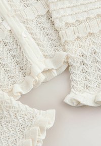 Cardigan en maille blanche avec des motifs délicats en dentelle, tissu texturé, bords volantés et boutons, présentant une apparence douce et légère.