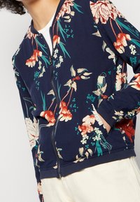 Veste zippée marine avec poignets et ourlet élastiques, présentant des motifs floraux et d'oiseaux rouges, crème et turquoise, portée avec un pantalon blanc.