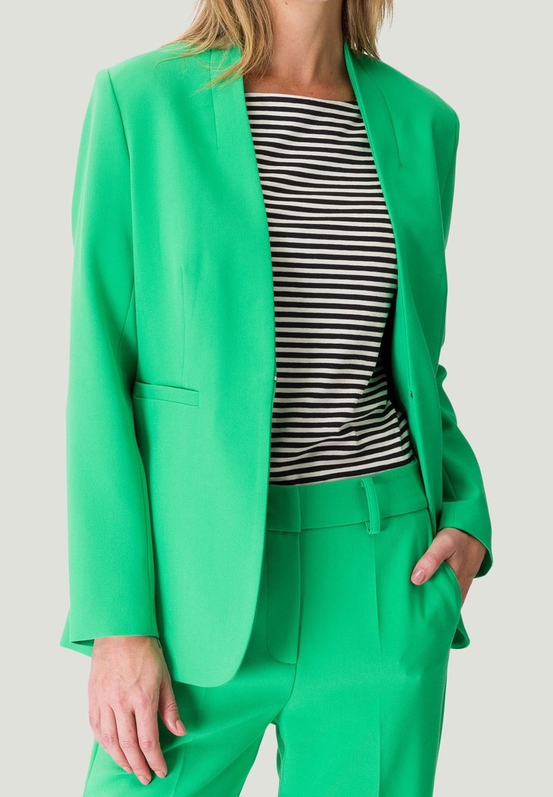 Femme portant un blazer vert vif et un pantalon assorti avec un haut rayé noir et blanc, debout avec une main dans la poche.
