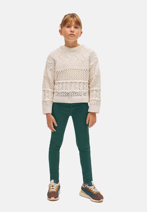 Suéter de punto color crema claro con patrones texturizados, combinado con pantalones verdes ajustados y zapatillas multicolor. Diseño simple y casual.