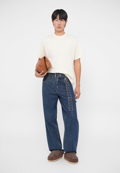 T-shirt beige clair à manches courtes, jeans bleu marine à taille large avec une ceinture accentuée de perforations, sac en cuir texturé marron et chaussures slip-on marron.