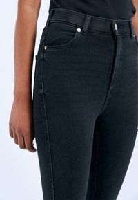 Black high-waisted denim jeans with a close fit, featuring front pockets, a zip fly, and metal button closure. Smooth texture and minimal stitching details.