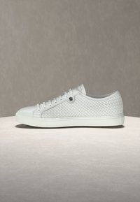 Baskets basses blanches avec côtés en cuir tissé, lacets à l'avant et semelle en caoutchouc plate, posées sur une surface et un fond neutres.