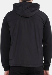 Giacca nera con cappuccio, caratterizzata da un rivestimento esterno liscio e maniche a maglia. Design semplice senza motivi visibili o elementi in metallo.