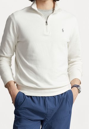 Man draagt een witte pullover met een kwart ritssluiting over een grijs hemd, blauwe broek met handen in de zakken en een horloge aan zijn linkerpols.