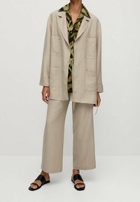 Beige lightweight jacket with large pockets over a green patterned shirt, paired with wide beige trousers and black sandals.