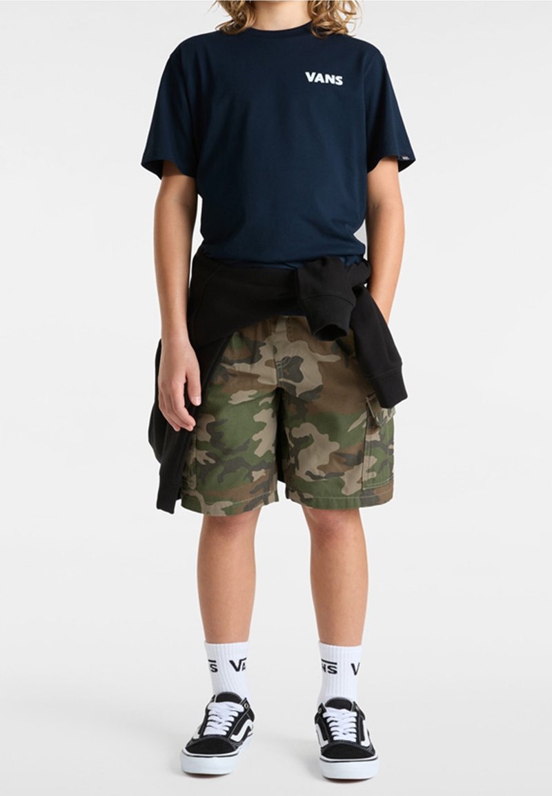 Jeune portant un t-shirt Vans bleu marine, un sweat noir attaché à la taille, un short camouflage, des chaussettes blanches Vans et des baskets noires Vans debout.