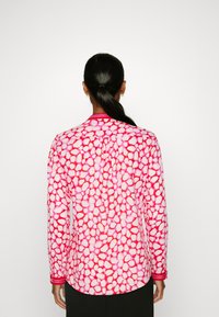 Camicia a maniche lunghe di colore rosa con motivo a pois. Presenta un orlo arrotondato e colletto a contrasto. Realizzata in tessuto leggero.