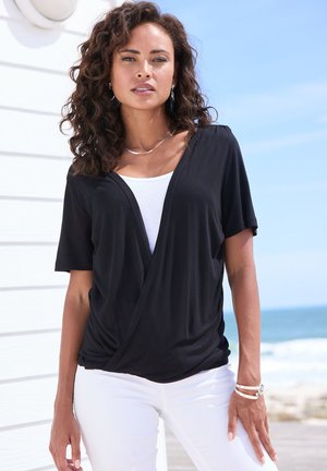 Woman with curly hair wearing black wrap top over white shirt and white pants, standing outdoors near beach and white wall.