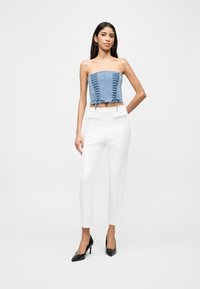 Γυναίκα με ανοιχτό μπλε κορσέ χωρίς τιράντες, λευκό ψηλόμεσο παντελόνι με ραφές και μαύρες γόβες με μυτερή μύτη, που στέκεται μπροστά σε λευκό φόντο.