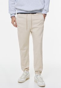 Pantaloni jogger beige chiaro con orli elasticizzati, vita con cordoncino e tasche laterali, abbinati a scarpe da ginnastica bianche con accenti leggeri.