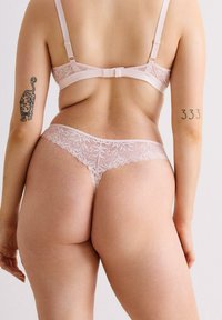 Spitzenunterwäsche-Set in Hellrosa mit floralem Muster, verstellbaren Trägern und einem Tanga-Schnitt, der eine glatte Textur betont.