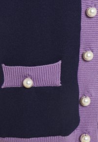 Cardigan bleu marine et lavande avec un panneau texturé, présentant des boutons semblables à des perles et une poche à rabat, alliant des tissus en maille lisse et côtelée.