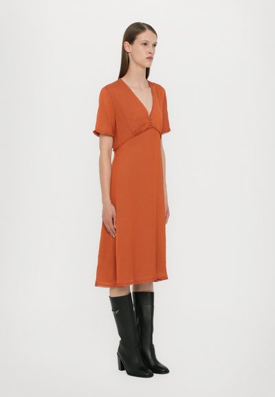 Vestido naranja a la altura de la rodilla con mangas cortas y escote en V, con un detalle de botones en la cintura. Combinado con botas altas negras.