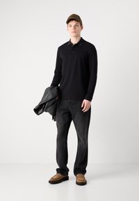 Μαύρο πουκάμισο polo με μακριά μανίκια, σκούρα τζιν, καφέ μοκασίνια και καφέ καπέλο. Κρατάει μια μαύρη δερμάτινη ζακέτα με απαλή υφή.