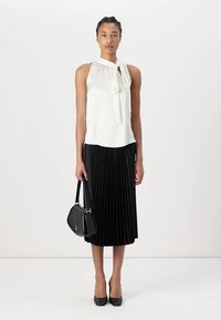 Blouse blanche sans manches avec un nœud au cou, associée à une jupe midi plissée noire. Le modèle tient un petit sac à main noir et porte des talons noirs.