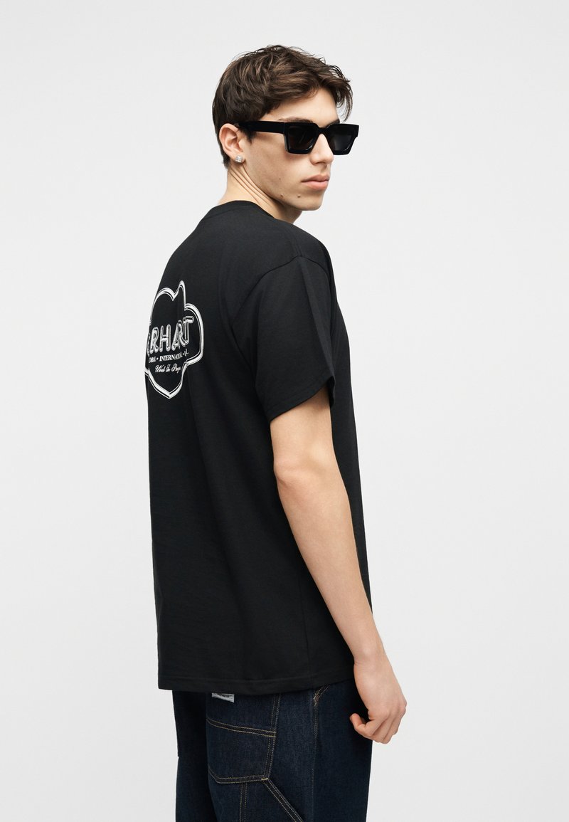 Giovane uomo con occhiali da sole neri e una T-shirt grafica nera con logo bianco sul retro, abbinata a jeans blu scuro, visto di profilo.