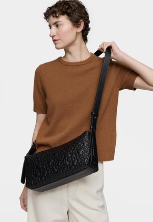 Donna che indossa un maglione marrone a maniche corte e pantaloni bianchi, che tiene una borsa a tracolla nera con trama e cinturino regolabile su sfondo bianco.