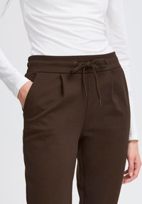 Naine kannab tumesarja paelaga pükse ja valget pika varrukaga pluusi, hoides ühte kätt taskus, taustal on lihtne taust.