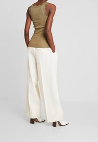 Woman wearing a beige ribbed tank top with lace trim, wide-leg white trousers, and white heeled boots, standing with hands in pockets.