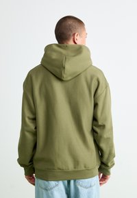 Person med kort hår iført en olivengrøn hoodie og lyseblå jeans, vist fra ryggen mod en ensfarvet hvid baggrund.