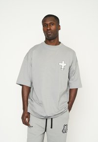 Camiseta gris de gran tamaño con mangas cortas, que presenta un diseño de cruz en blanco y negro en el pecho. Hecha de material de algodón suave.