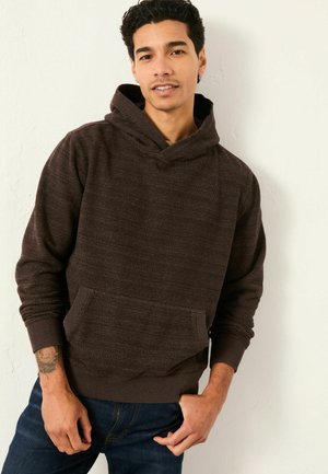 Mann mit dunklem lockigem Haar, der einen braunen Kapuzenpullover und dunkle Jeans trägt, steht vor einer hellen, strukturierten Wand.