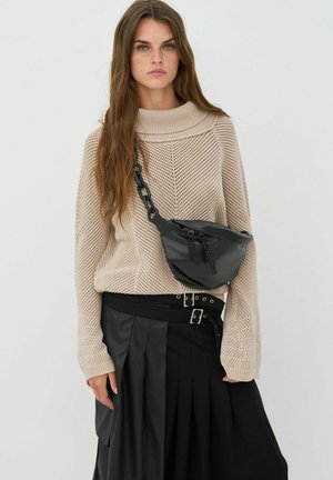 Žena s dlhými hnedými vlasmi, oblečená v béžovom pletenom svetri, čiernej plisovanej sukni s opaskom a čiernej kabelke cez rameno s veľkým reťazovým remienkom.