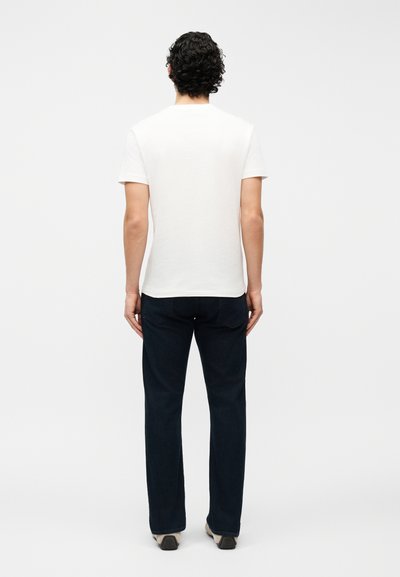 Άνδρας με σγουρά σκούρα μαλλιά που φοράει απλό λευκό κοντομάνικο μπλουζάκι και σκούρα τζιν παντελόνια, στέκεται με την πλάτη στραμμένη σε λευκό φόντο.