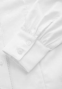 Polsino di camicia bianca con dettagli plissettati e due bottoni, realizzato in tessuto liscio con un delicato bordo in pizzo lungo il margine.