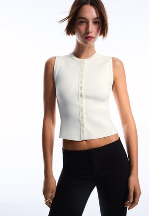 Młoda kobieta o krótkich brązowych włosach, ubrana w białą, bezrękawnikową bluzkę na guziki oraz czarne spodnie z wysokim stanem, stoi na tle jednolitej, białej ściany.