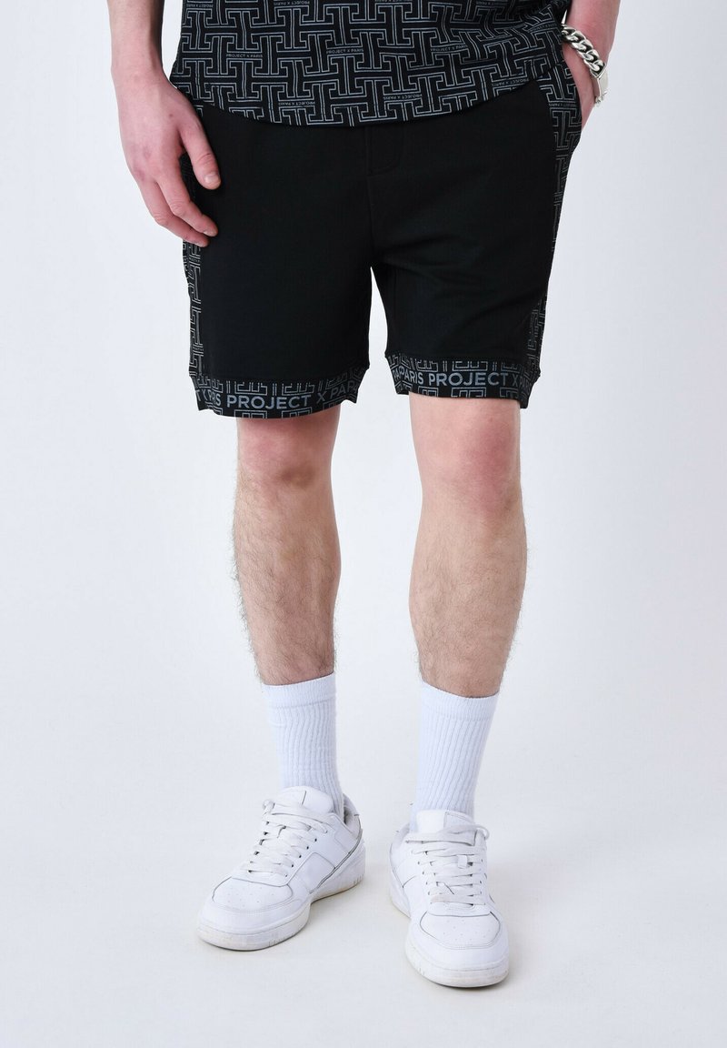Shorts noirs avec un ourlet à motif portant le texte "PROJECT". Associés à des chaussettes blanches et des baskets, mettant en avant un look décontracté et sportif.