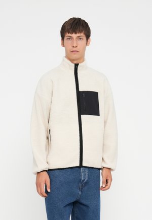 Fleece jakke i creme med en sort lynlåslomme på brystet. Har en høj krave og lynlås-lukning, samt elastiske ærmer og en afslappet pasform.