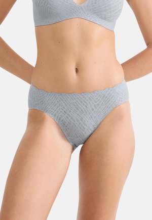 Femme portant un soutien-gorge et une culotte assortis gris clair sans couture avec un motif géométrique subtil, debout les mains sur les hanches sur un fond blanc.