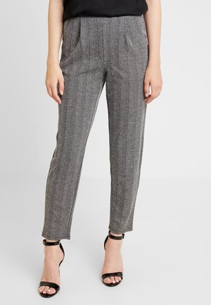 Femme vêtue d'un pantalon droit en chevrons gris avec poches latérales et sandales à talons hauts noires à bride cheville, debout devant un fond blanc.
