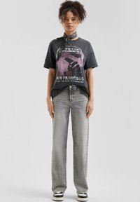 Grafische T-shirt in vervaagd zwart met paarse en witte print; ontspannen pasvorm. Gecombineerd met rechtzittende, lichtgrijze jeans en zwarte sneakers.