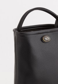 Bolso de mano negro con material sintético texturizado, asas redondeadas y un botón dorado en la parte frontal. Diseño compacto adecuado para el uso diario.