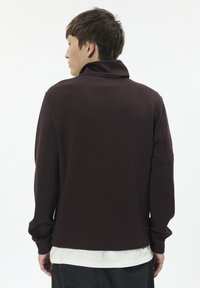 Dunkelbrauner Pullover mit hohem Kragen, langen Ärmeln und lockerer Passform. Hergestellt aus weichem Stoff mit glatter Textur und minimalen Nähten.