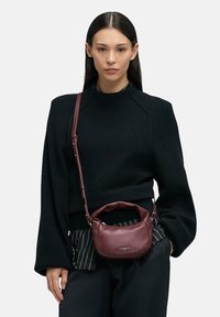 Czarny dziany sweter z długimi rękawami, zestawiony z pasiastymi spodniami. Trzyma małą bordową skórzaną torebkę z zaokrąglonym uchwytem.