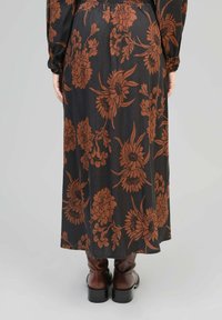 Robe maxi à motif floral en tissu noir avec des fleurs brunes, dotée d'une taille smockée et de manches longues, associée à des bottes à talons brunes.