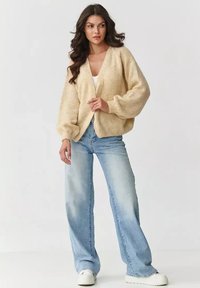 Jasno beżowy sweter z dzianiny z balonowymi rękawami, w dekolcie w serek, noszony na białą bluzkę, w połączeniu z jasnoniebieskimi spodniami typu wide-leg i białymi trampkami.