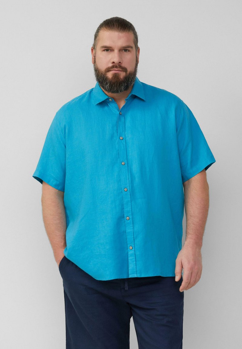 Kortebladige, knoopshirt in fel turquoise linnenstof. Heeft een klassieke kraag en een subtiele glans, gecombineerd met donkere broeken.